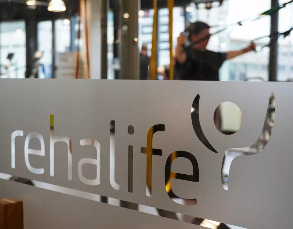 Blick durch eine Scheibe im Rehazentrum. Auf der Scheibe ist ein Streifen Milchglas-Folie aufgebracht, aus der das Logo von rehalife ausgeschnitten wurde. Im Hintergrund sind man verschwommen Menschen an Geräten trainieren.