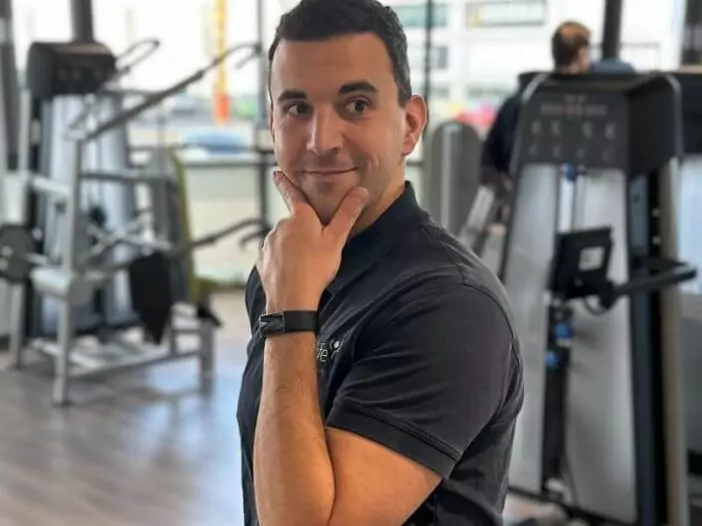Ein Mitarbeiter posiert lächelnd für die Kamera. Er trägt ein dunkelgraues Poloshirt mit Logo auf der Brust. Im Hintergrund sind Sportgeräte und eine trainierende Person zu sehen.
