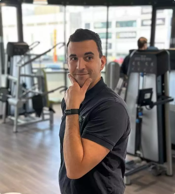 Ein Mitarbeiter posiert lächelnd für die Kamera. Er trägt ein dunkelgraues Poloshirt mit Logo auf der Brust. Im Hintergrund sind Sportgeräte und eine trainierende Person zu sehen.