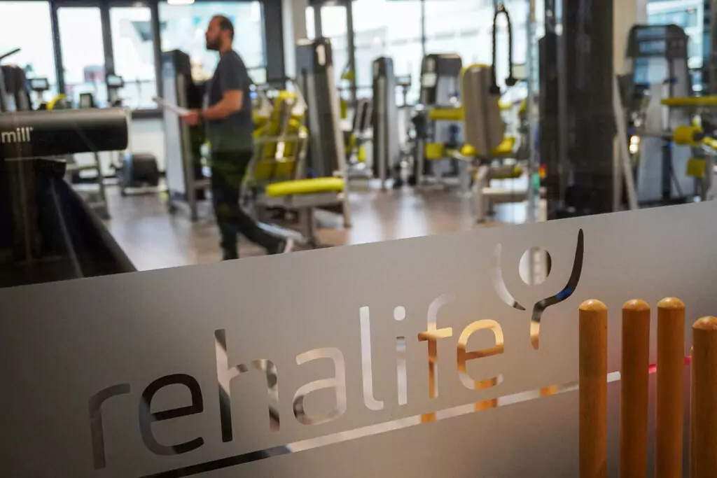 Blick durch eine Scheibe in den Kraftraum. Auf der Scheibe ist ein Streifen Milchglas-Folie aufgebracht, aus der das Logo von rehalife ausgeschnitten wurde. Im Hintergrund sind man verschwommen die Geräte und einen Trainer.