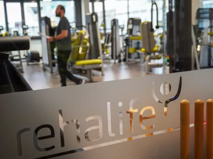 Blick durch eine Scheibe in den Kraftraum. Auf der Scheibe ist ein Streifen Milchglas-Folie aufgebracht, aus der das Logo von rehalife ausgeschnitten wurde. Im Hintergrund sind man verschwommen die Geräte und einen Trainer.