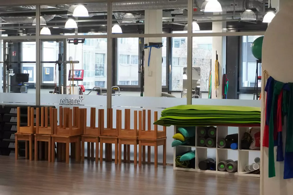Ansicht eines Raumes im Rehazentrum. Man sieht eine Glasfront, die den Raum zum Kraftraum trennt. An der Front stehen Hocker und ein Regal mit Faszienrollen und Sportmatten.