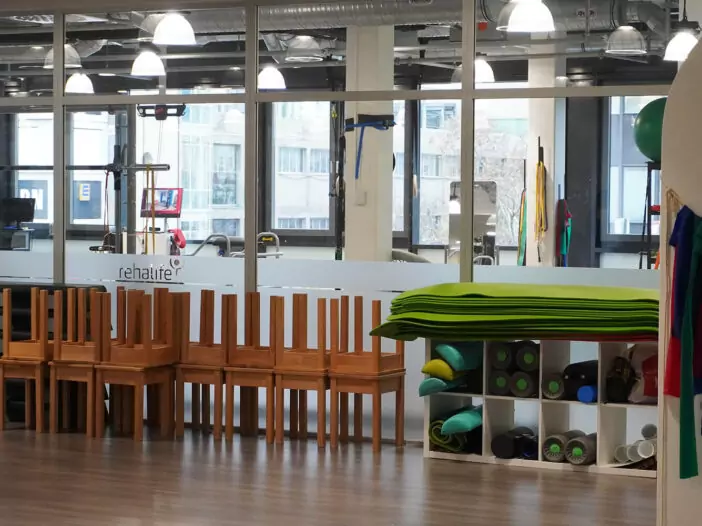 Ansicht eines Raumes im Rehazentrum. Man sieht eine Glasfront, die den Raum zum Kraftraum trennt. An der Front stehen Hocker und ein Regal mit Faszienrollen und Sportmatten.