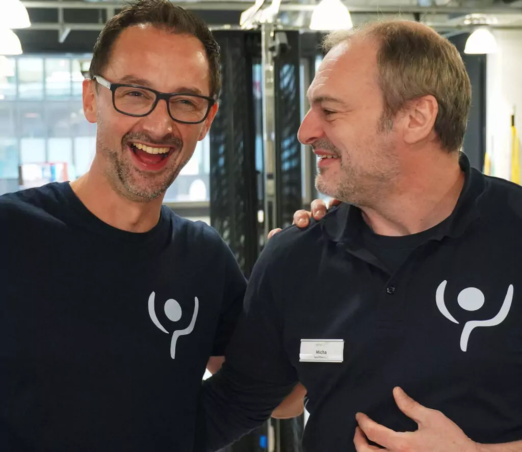 Zwei Mitarbeiter lachen. Sie tragen beide ein dunkelblaues T-Shirt mit der Bildmarke von rehalife.