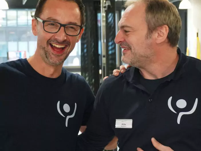 Zwei Mitarbeiter lachen. Sie tragen beide ein dunkelblaues T-Shirt mit der Bildmarke von rehalife.