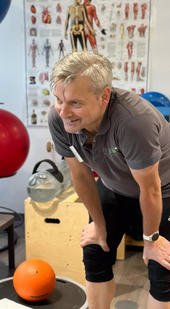 Ein Mitarbeiter mit grauem Poloshirt und Logo auf der Brust stützt sich lächelnd auf seine Knie. Im Hintergrund sieht man ein großes Plakat mit der Anatomie eines Menschens und zahlreiche Gymnastikbälle und andere Geräte.