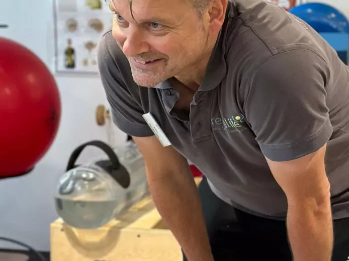 Ein Mitarbeiter mit grauem Poloshirt und Logo auf der Brust stützt sich lächelnd auf seine Knie. Im Hintergrund sieht man ein großes Plakat mit der Anatomie eines Menschens und zahlreiche Gymnastikbälle und andere Geräte.