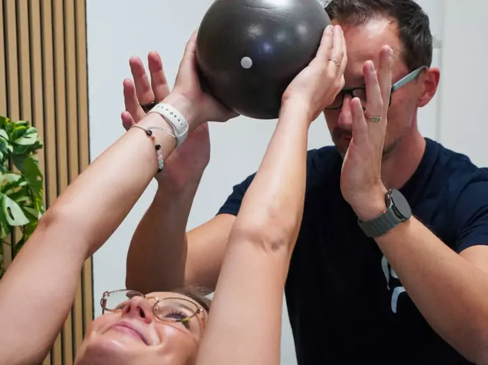 Eine junge Frau liegt auf dem Rücken, man sieht ihr Gesicht und ihre Arme, die sie nach üben über ihren Kopf streckt. Sie hält einen schwarzen Gummiball in der Hand. Hinter ihr sitzt am Kopfende ein Therapeut der die Hände in der Nähe des Balles hält und sie anleitet.