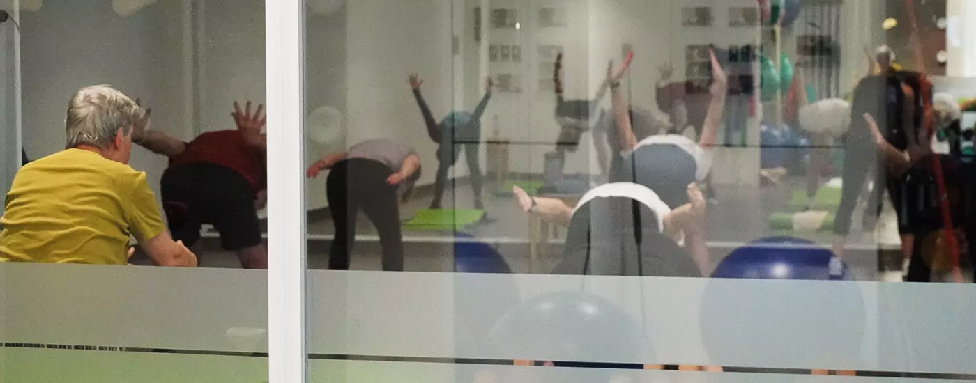 Blick durch eine Glasscheibe. Man sieht eine Gruppe von Kursteilnehmern bei dem Ausüben einer Übung. Sie beugen sich nach vorne und strecken die Arme nach hinten und oben.