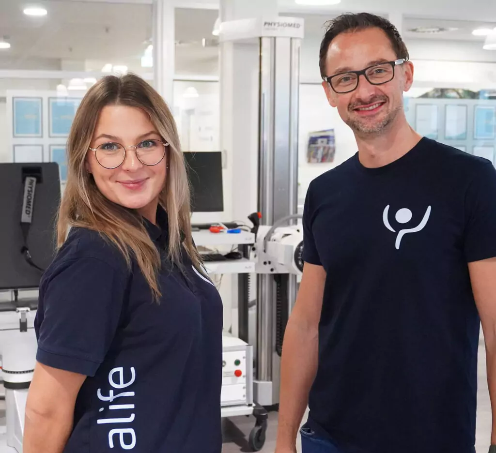 Links eine junge Mitarbeiterin, rechts ein Mitarbeiter, beide in dunkelblauen T-Shirts mit weißem rehalife-Logo. Sie lächeln in die Kamera. Im Hintergrund stehen einige Geräte.