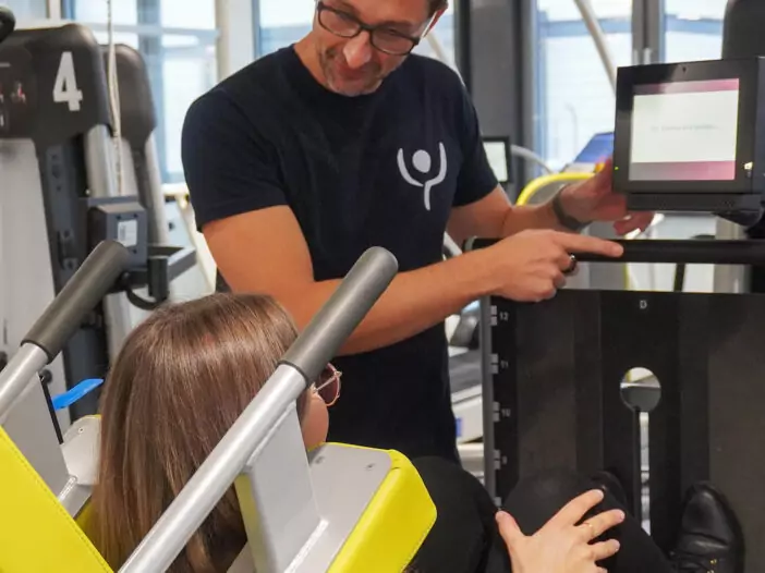 Eine junge Frau sitzt auf einem Gerät, mit dem Man die Beine trainiert. Die Beine sind angewinkelt. Ein Mitarbeiter steht neben ihr und erklärt die Benutzung des Gerätes.