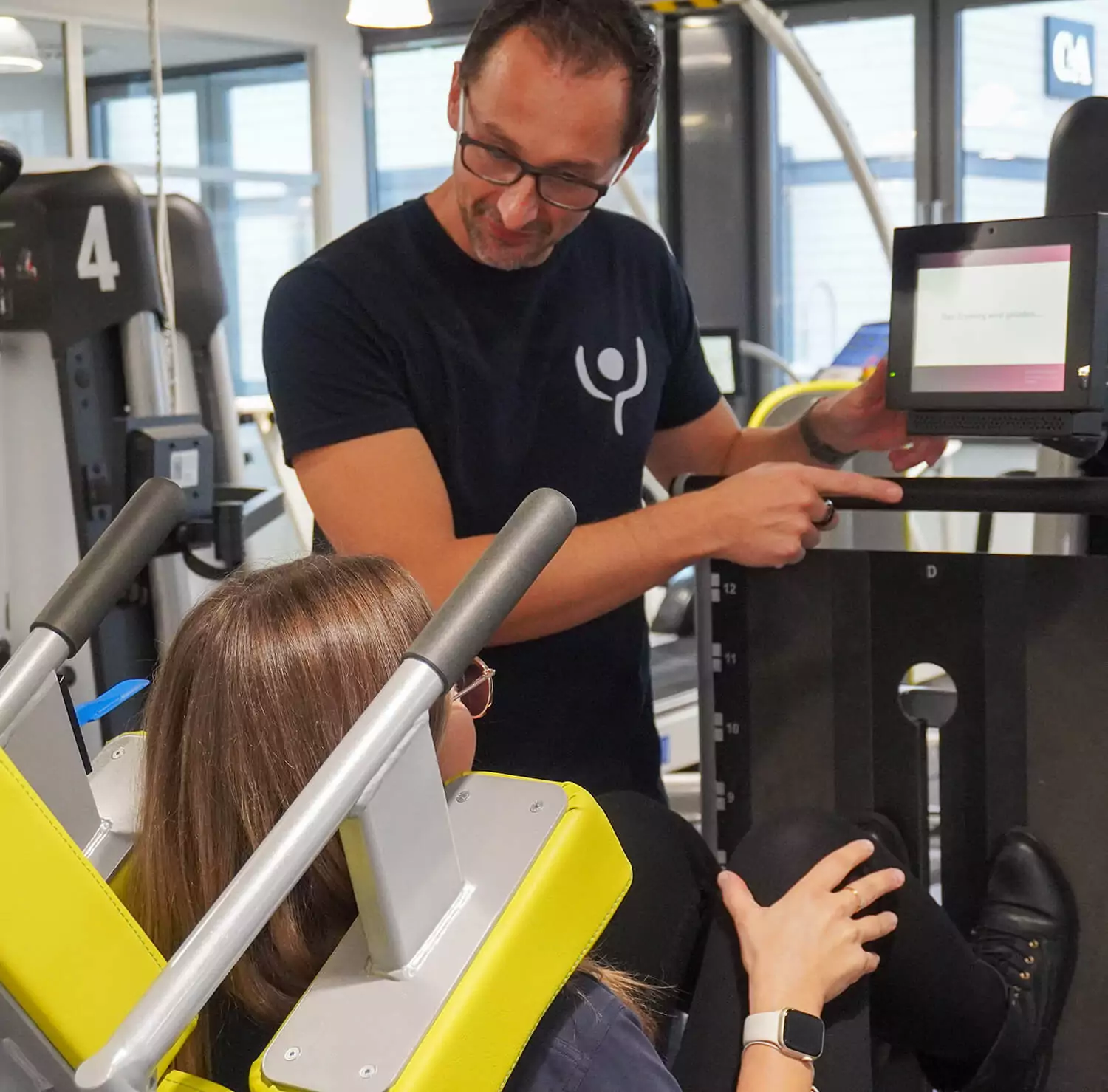 Eine junge Frau sitzt auf einem Gerät, mit dem Man die Beine trainiert. Die Beine sind angewinkelt. Ein Mitarbeiter steht neben ihr und erklärt die Benutzung des Gerätes.