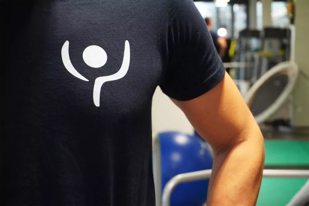 Ausschnitt einer männlichen Person mit einem dunkelblauen T-Shirt. Darauf ist. die Bildmarke von rehalife zu sehen. Eine schematische Darstellung einer Person, die die Arme hochstreckt.