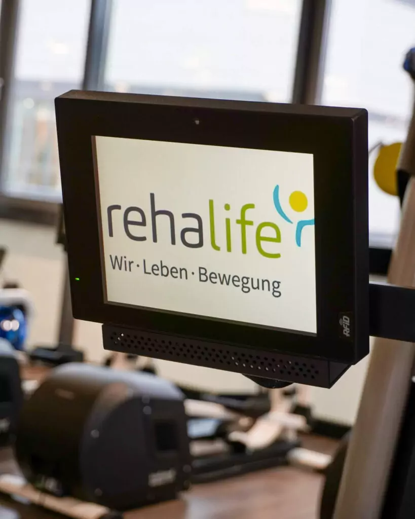 Ein Monitor mit dem Logo von rehalife. Er gehört zu einem der Sportgeräte. Im Hintergrund sieht man weitere Geräte und die Fensterfront der Praxis