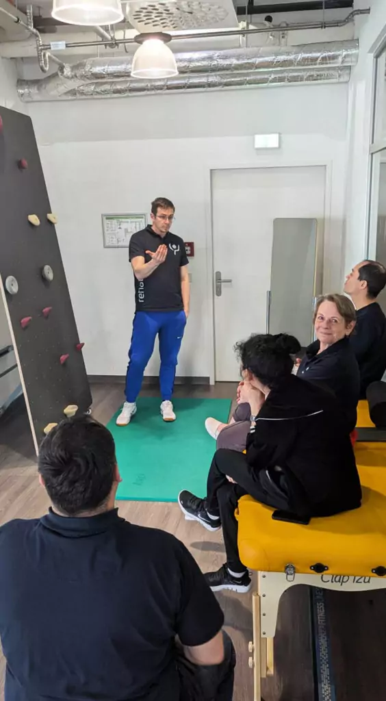 Fünf Personen in einem Praxisraum. Ein Mann steht, die anderen sitzen vor ihm. Er erklärt gerade die Benutzung einer Kletterwand, neben der er steht. Eine der sitzenden Frauen schaut in die Kamera und lächelt.